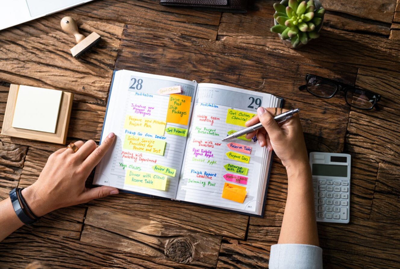 Agenda aperta su una scrivania in legno con appunti colorati e post-it per la gestione del tempo e la pianificazione del lavoro aziendale.