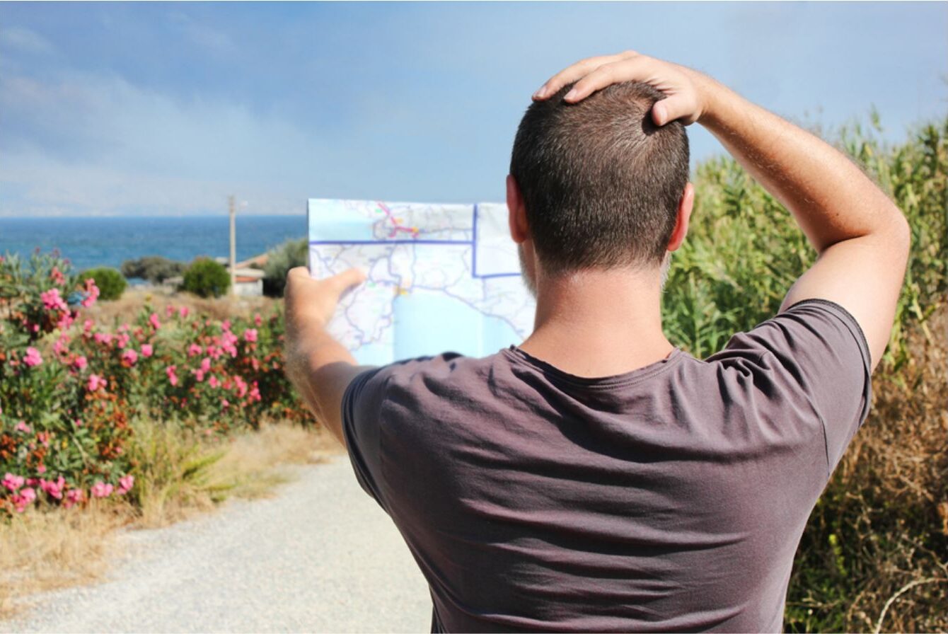 Turista di spalle confuso mentre consulta una mappa cartacea su un sentiero panoramico, cercando indicazioni per raggiungere una caletta al mare.